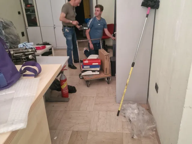 Due componenti del team riordinano l'ufficio della sede, con faldoni e uno scatolone su un carrello