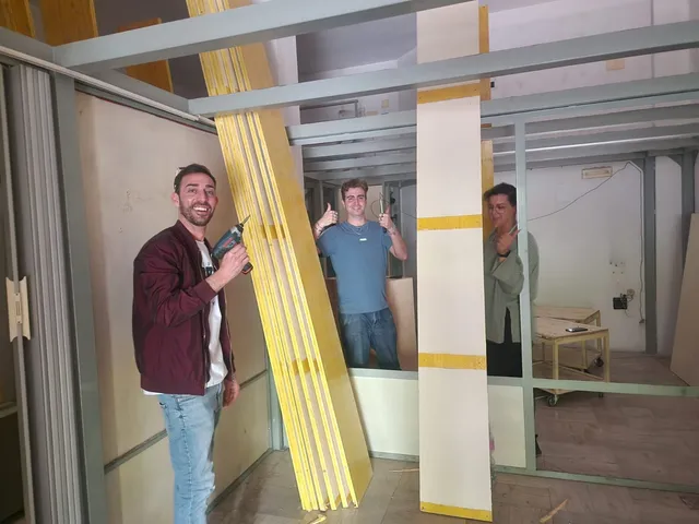 Tre membri del team, con il pollice alzato, tra le assi di legno della parete in costruzione